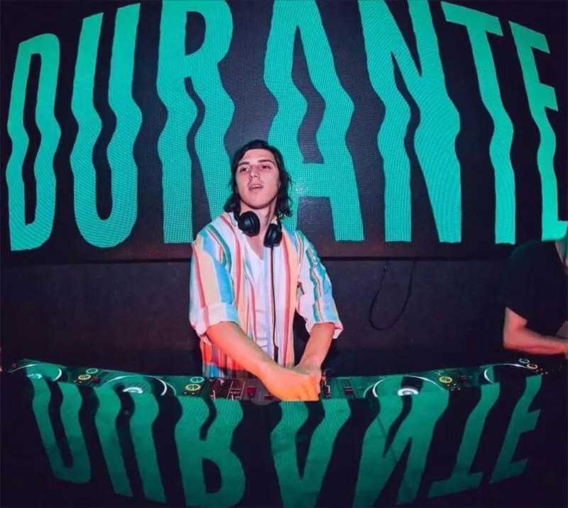 Durante DJing in a black booth, with his name "Durante" in teal green and in caps projected on the wall behind him