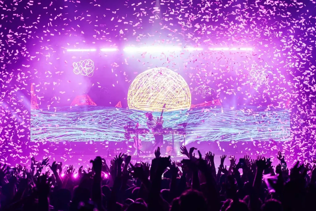 San Holo performing on stage illuminated in neon purple light with confetti falling