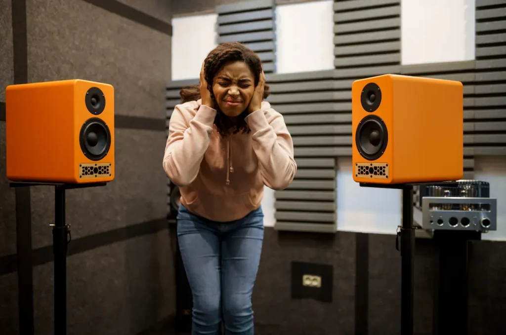 A woman standing between two orange speakers, covering her ears with her hands