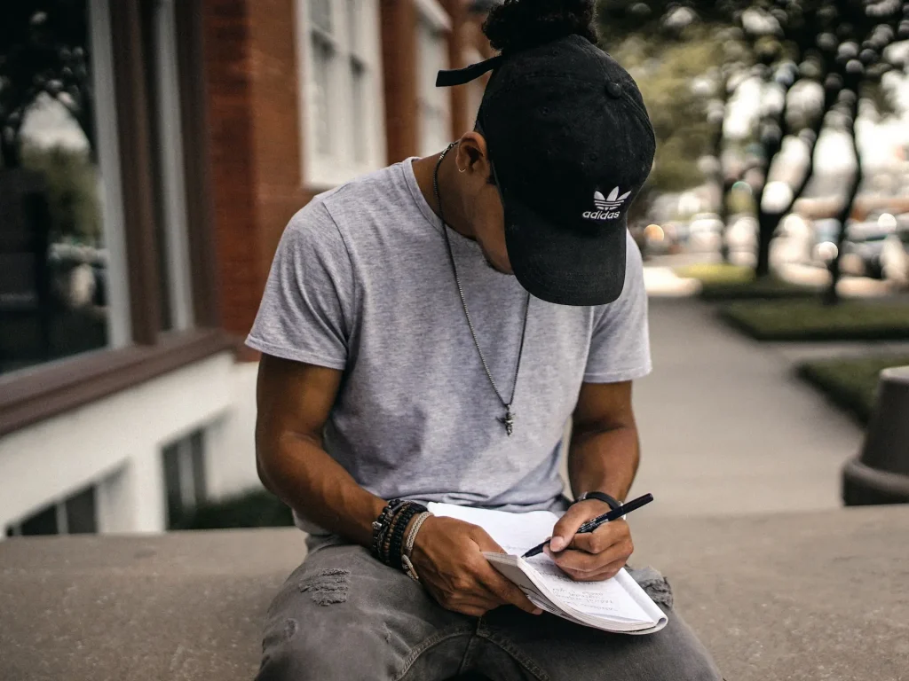 A man wearing a cap sitting outside and writing in a notebook