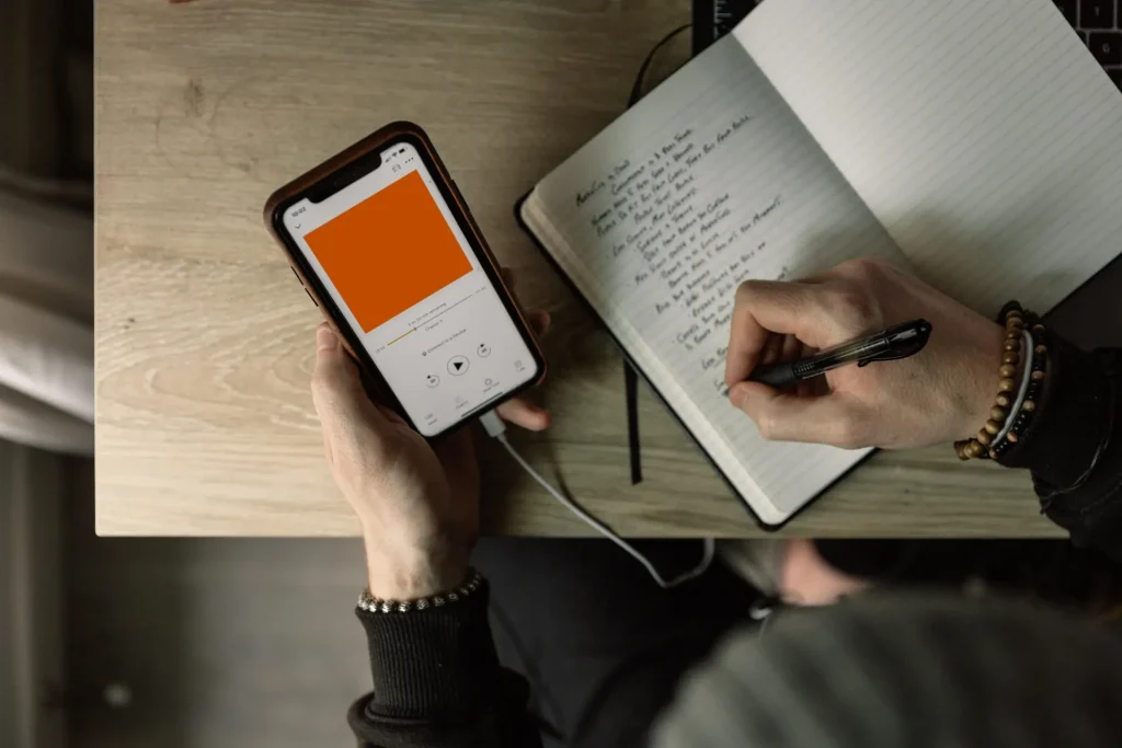A person writing in a notebook, holding a phone playing music in the other hand
