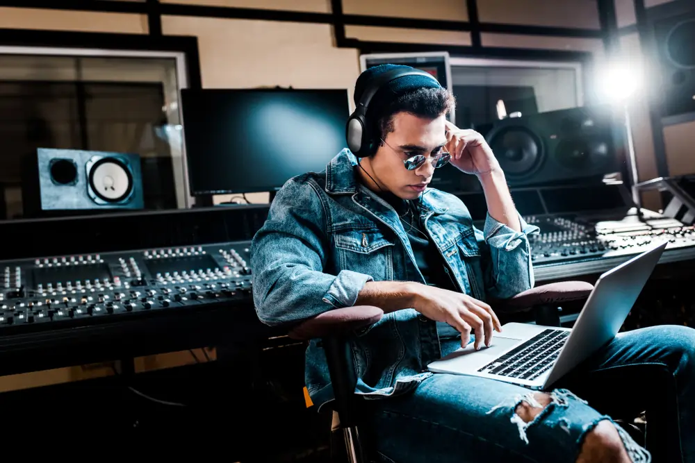 A man wearing headphones, sitting in a chair with a laptop on his lap in a studio