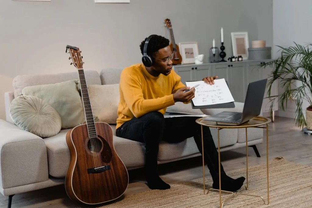 A man wearing headphones sitting on a couch next to an acoustic guitar showing sheet music to a laptop screen