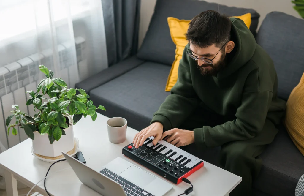 A man sitting on a couch in front of a laptop computer playing a MIDI keyboard