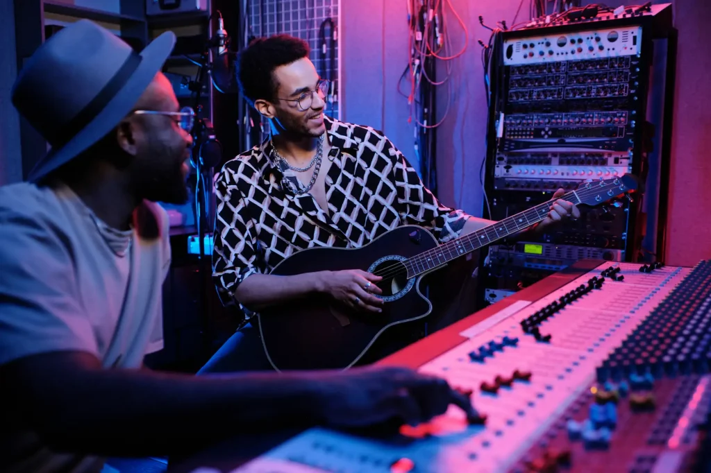 Two men in a recording studio. One man playing the guitar and the other adjusting levels on a mixure.